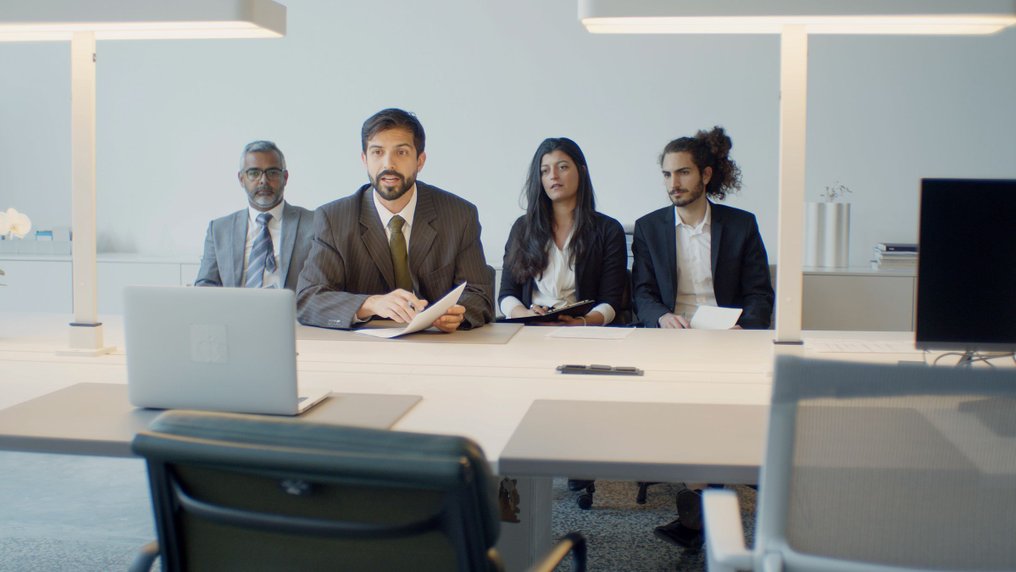 Quatre collègues assis côte à côte à une table de réunion suivent une visioconférence face à un écran
