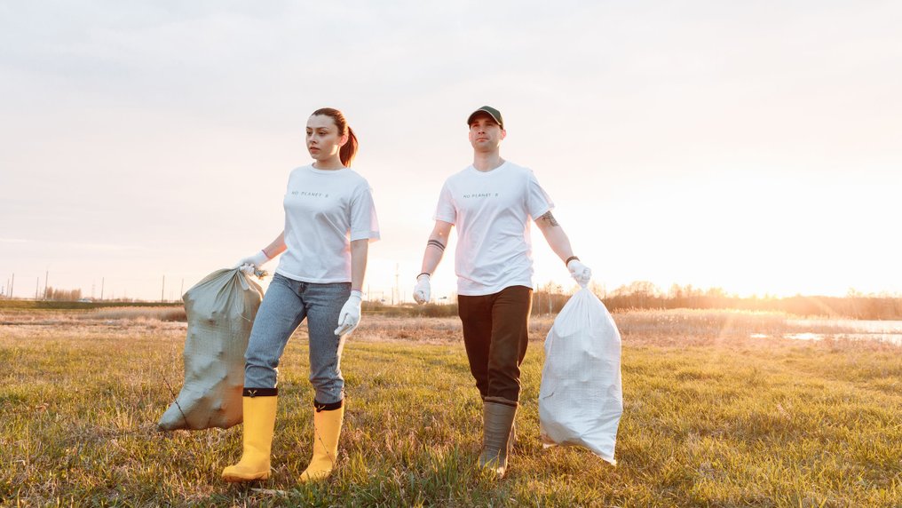 Deux bénévoles en bottes ramassent des déchets dans des sacs sur un terrain en friche, au coucher du soleil