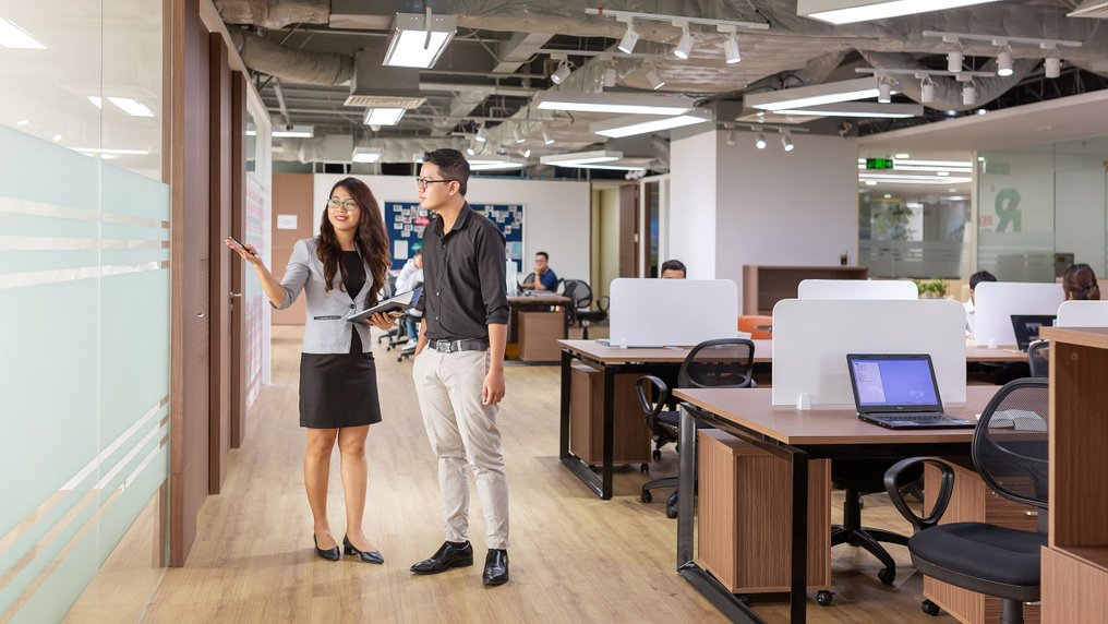 Dans un plateau de bureaux ouvert, une salariée présente les lieux à un collègue qui l’écoute
