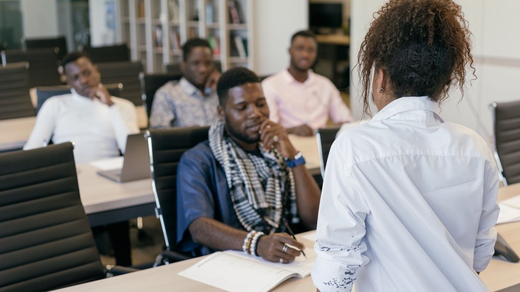Une formatrice, vue de dos, s’adresse à des stagiaires adultes assis à leurs tables dans une salle de formation