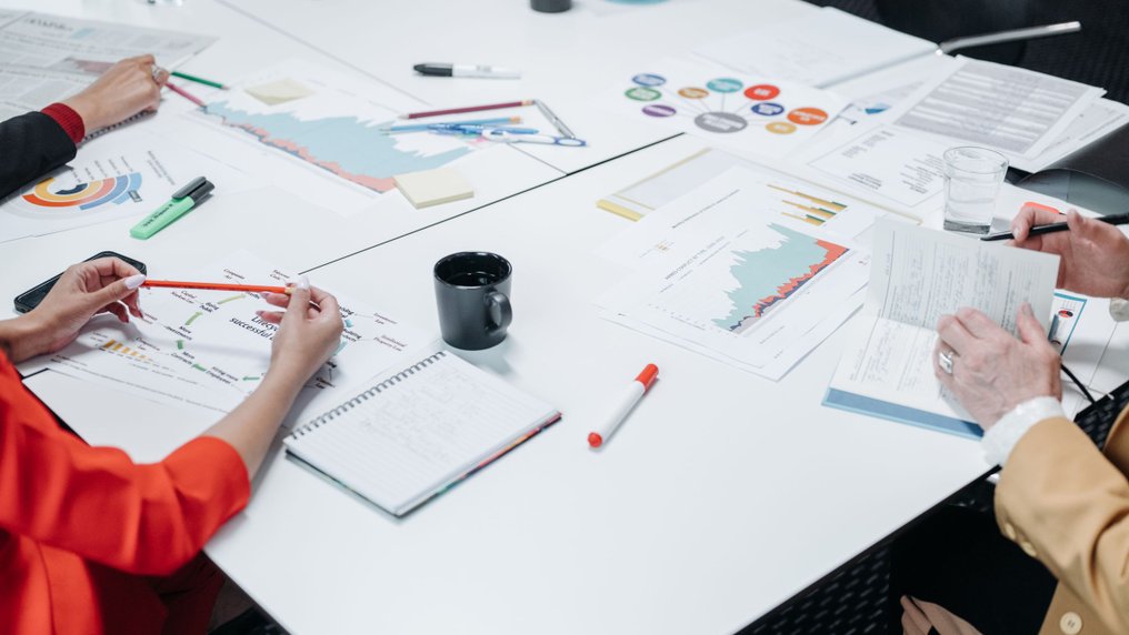 Autour d’une table de bureau, des collègues annotent des tableaux et des graphiques imprimés, avec des carnets et des tasses de café