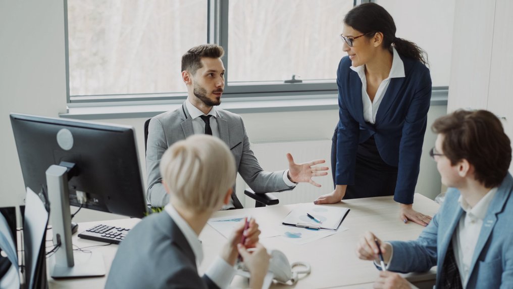 Debout près d’une table de bureau, une manager s’adresse à quatre collègues assis devant leurs ordinateurs, qui l’écoutent et lui répondent