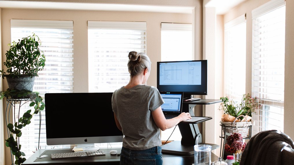 Dans un bureau lumineux garni de plantes, une salariée travaille debout à un bureau réglable, devant un écran et un ordinateur portable