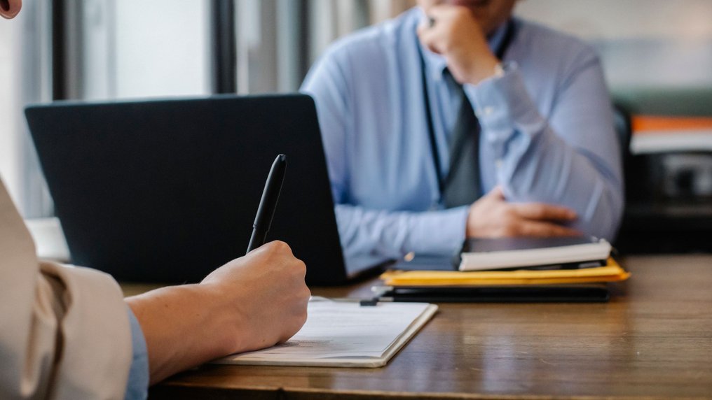 Entretien dans un bureau : une salariée prend des notes face à un responsable, un ordinateur portable et des documents sur la table