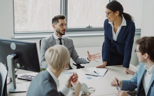 Debout près d’une table de bureau, une manager s’adresse à quatre collègues assis devant leurs ordinateurs, qui l’écoutent et lui répondent